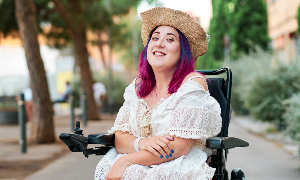 Image: Woman with colourful hairs, a sunhat and a dress sitting in a wheel chair and smiling in a garden.
