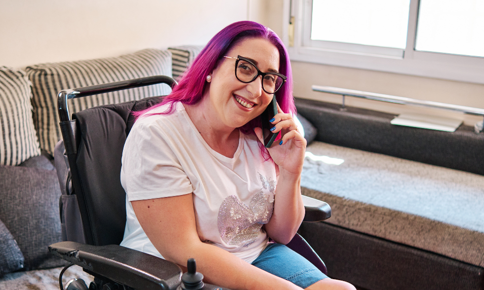 A woman in a pink shirt with brightly coloured hair who uses a wheelchair is making a call on her mobile phone