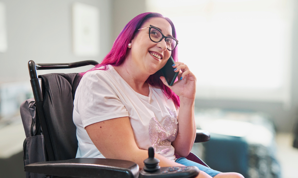 A woman in a pink shirt with brightly coloured hair and glasses who uses a wheelchair is on the phone calling Yooralla