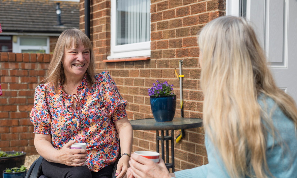 IMAGE: A client who uses a wheelchair is smiling and having a cup of tea outdoors with their Behaviour support practioner