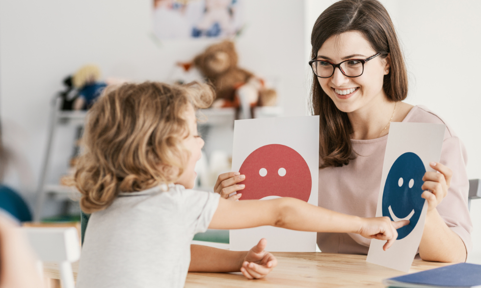 IMAGE: A Behaviour Support Practitioner is sitting with a young client holding up two cards, a smiley face and a frowning face