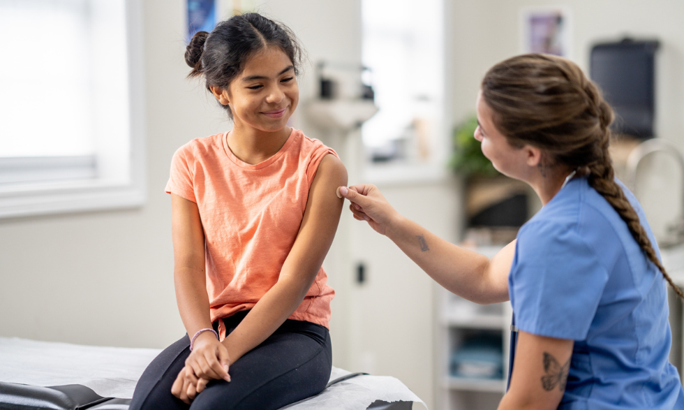 Image: Young girl in an an orange t-shirt receiving a vaccination from a nurse