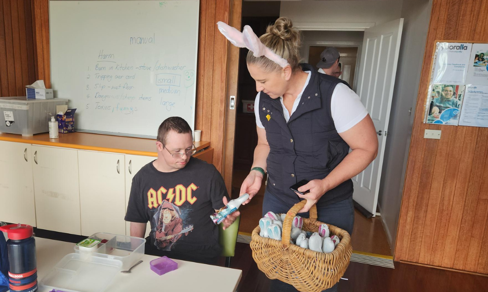 Image description: Bank staff member passing a chocolate to a Yooralla client. The staff member is wearing bunny ears and holding a basket of chocolate eggs.