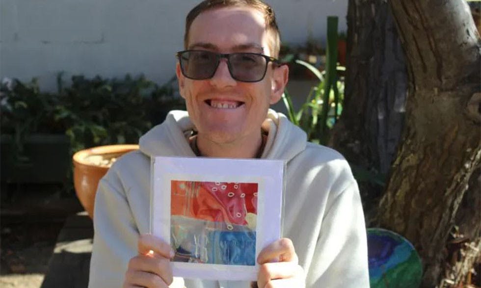 IMAGE: ben holding his intricate artwok up to the camera - Ben wears tinted sunglasses and a white hoodie and is sitting outside under a tree