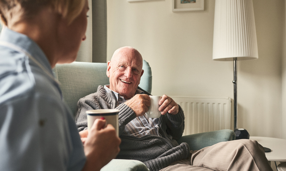 Image: Older man sitting, smiling and enjoying a cup of tea with his support worker