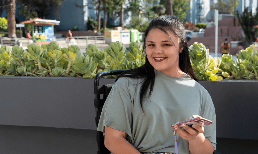 IMAGE: A young person who uses a wheelchair is sitting outside in the sun looking to camera with their phone in their hand