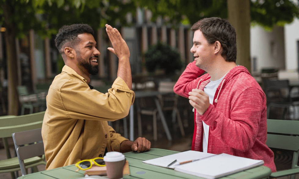 IMAGE: A person is sitting at a cafe with their support worker they are giving each other a high-five!