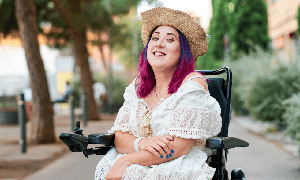 Image: Woman with colourful hairs, a sunhat and a dress sitting in a wheel chair and smiling in a garden.