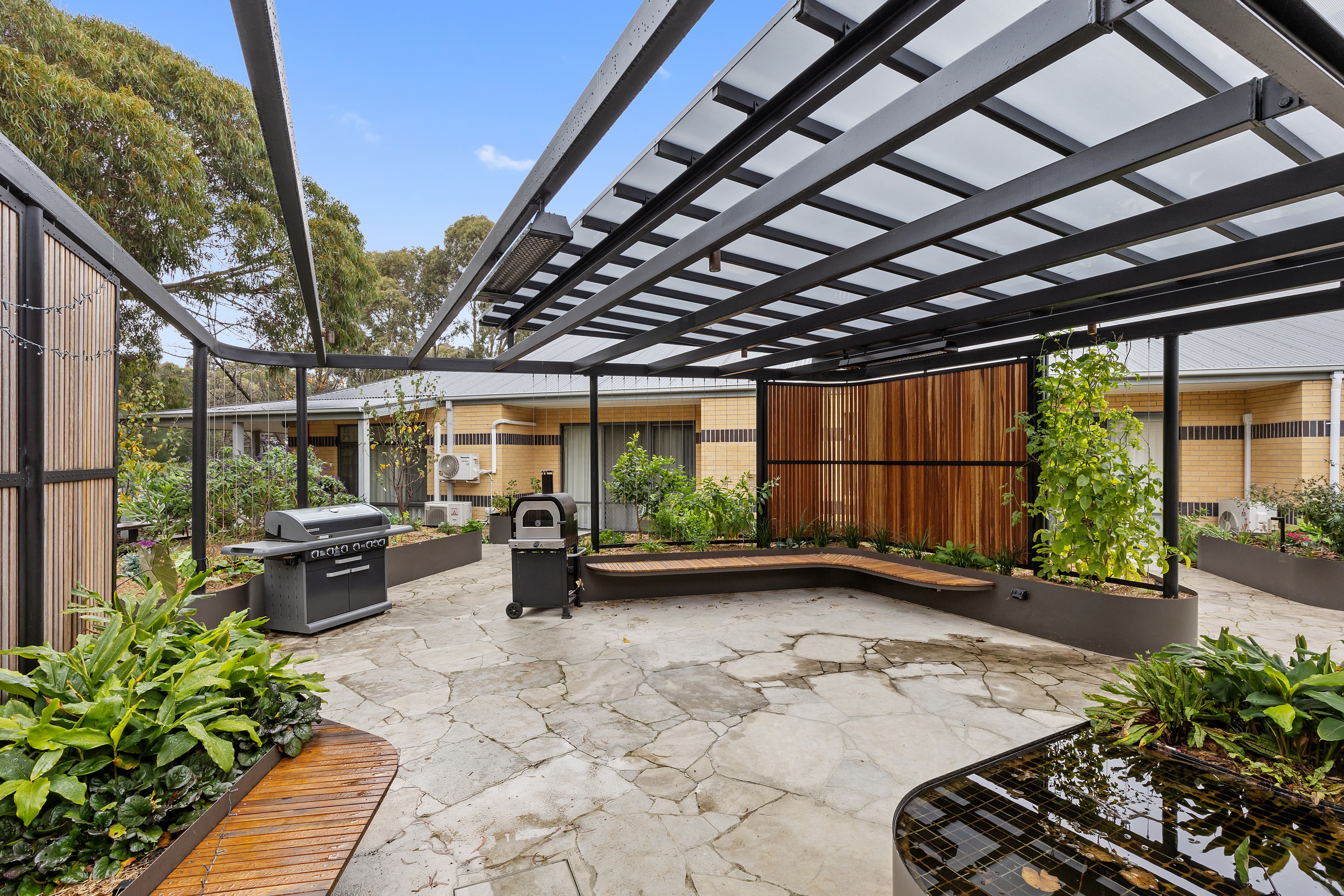 Image: Thornbury's Sensory Garden. A beautiful garden and blue skies with wooden benches, a pizza oven, a BBQ, concrete ground and healthy green plants.