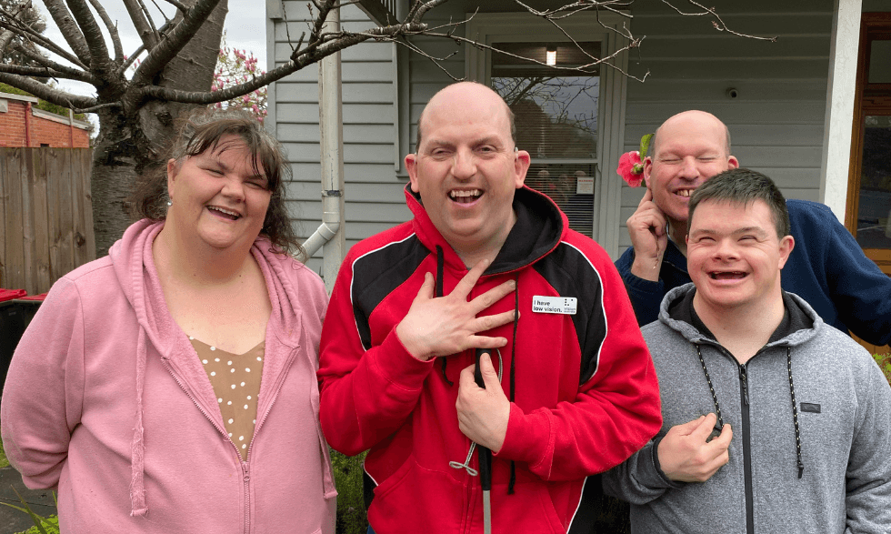 IMAGE: a group of clients are standing together smiling to camera, a client in the back holds a flower up to his ear posing for the camera