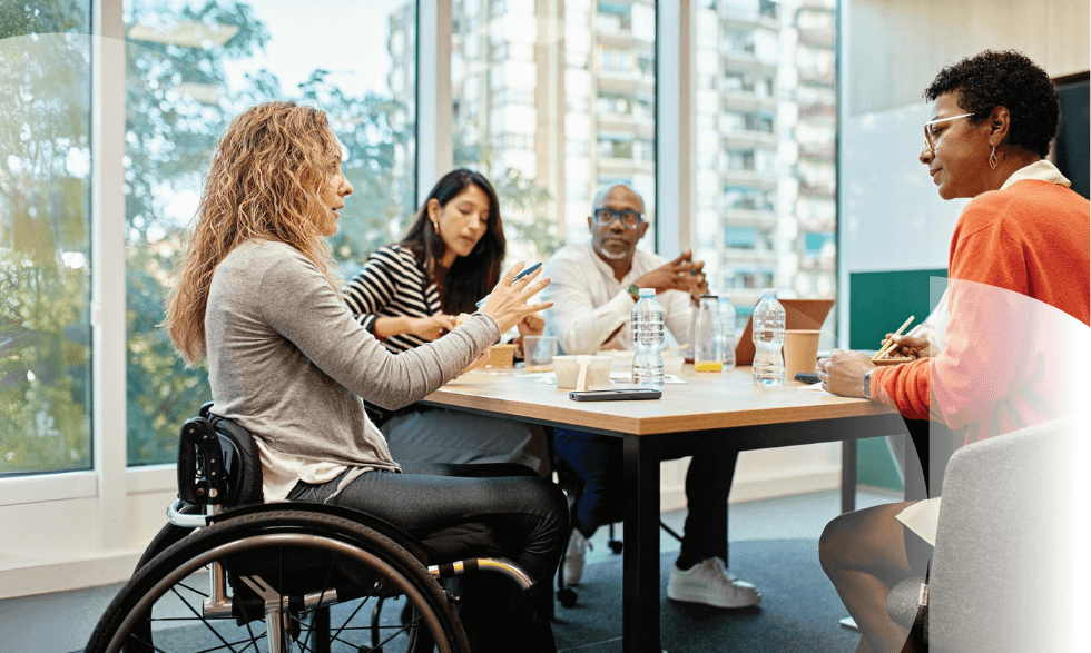 IMAGE: A person who uses a wheelchair is seated at a meeting table in a modern office explaining a concept to a group of colleagues