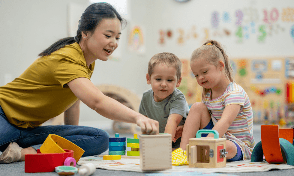 IMAGE: Two young children are sitting on the floor with their physiotherapist. They are working with wooden blocks.