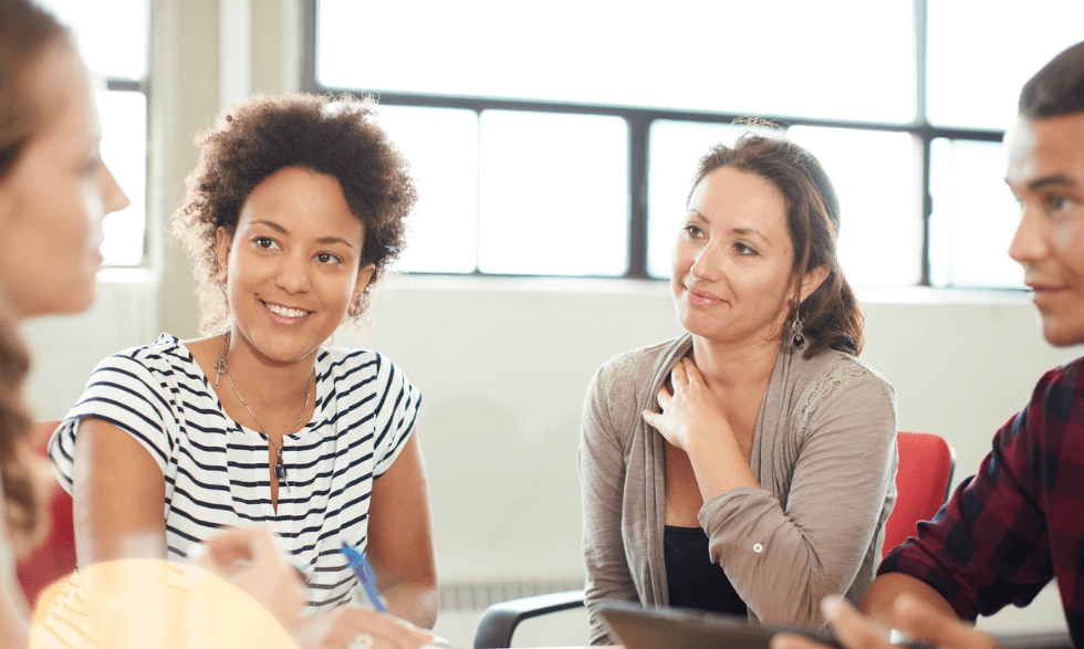 a group of four people sitting around an office style table, taking notes and chatting