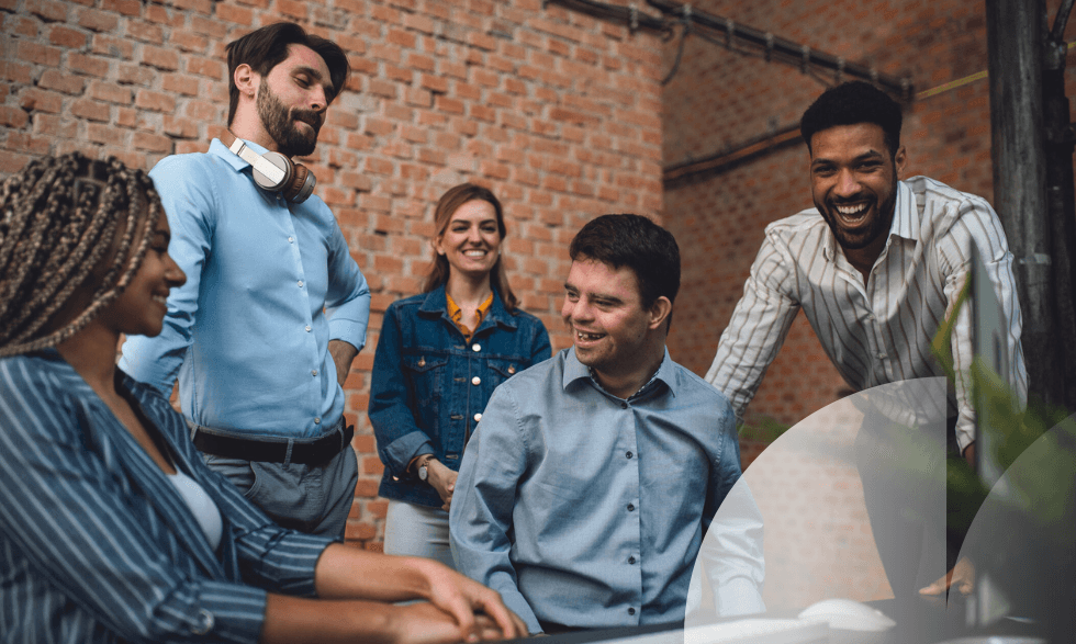 IMAGE: a group of people with diverse abilities sitting together in an office - a gentleman with disability in a button up blue shirt is at the centre, he is smiling and laughing