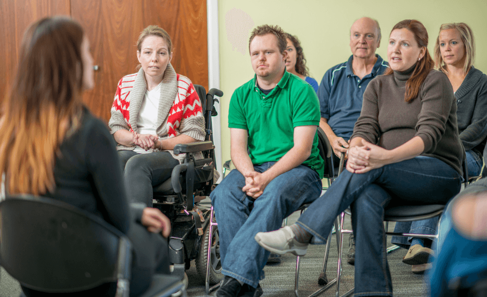 IMAGE: A group of people with disability are sitting in a room with a speaker discussing a serious topic