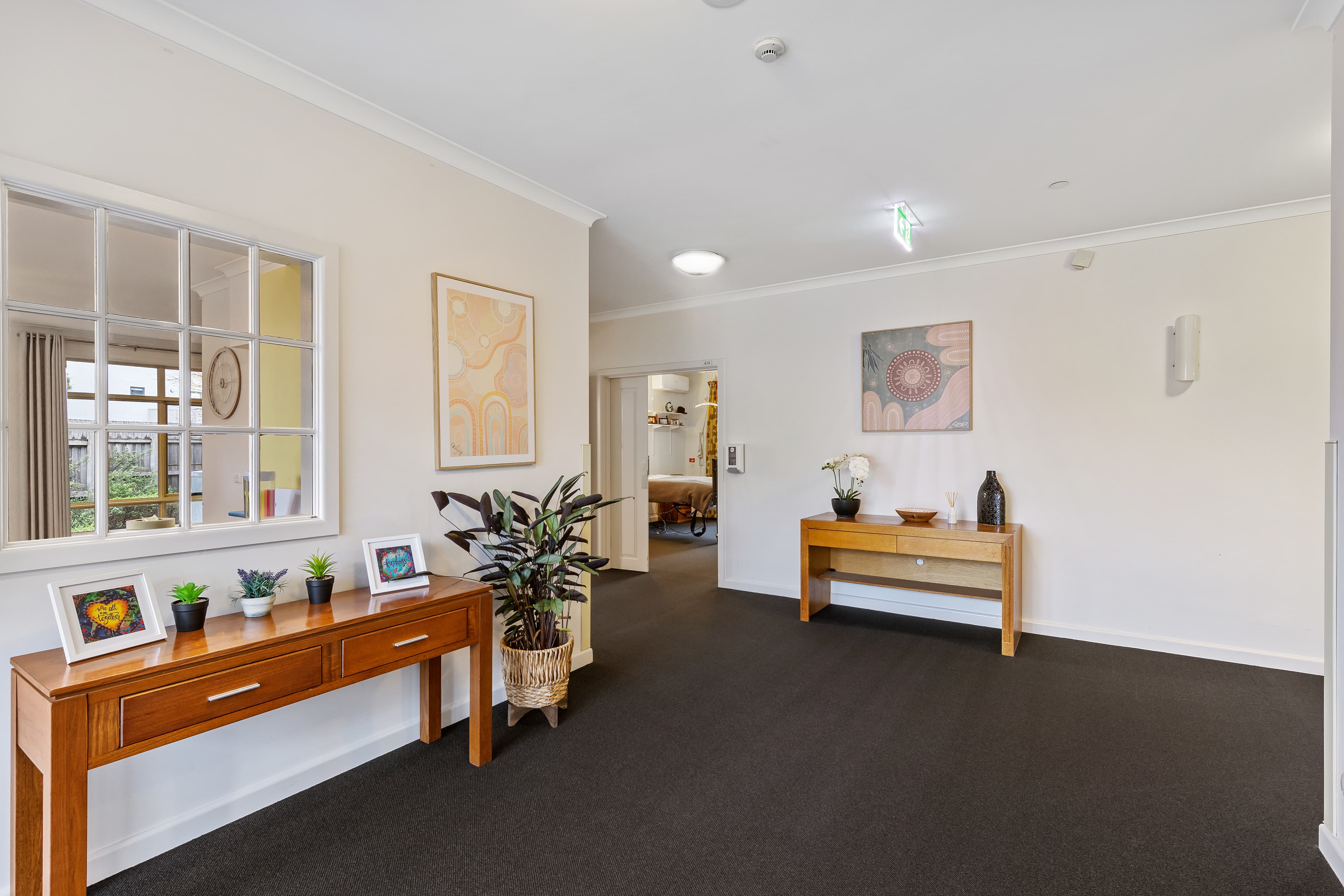 Image: An entry passage with a mirror on white walls and a painting to its right. A wooden sideboard with little cactuses and a fern to its right. The back wall also has a wooden side board with vases and plants on top of it,