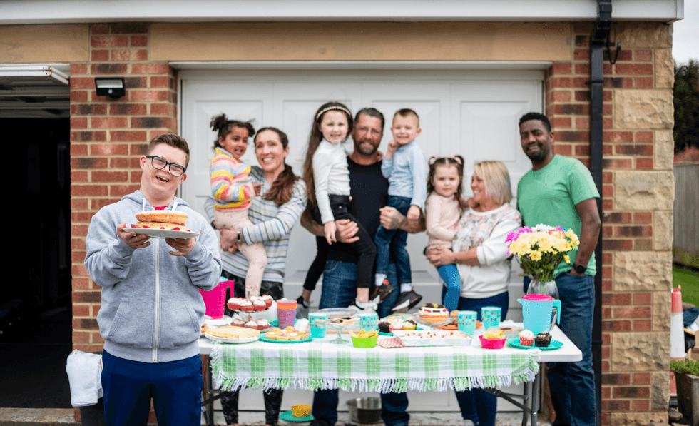 Image: Person with a disability holding a cake in front of group of adults and children at a bake sale